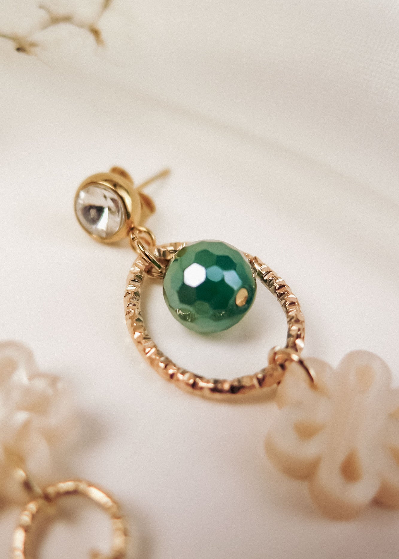 A close-up of a gold earring featuring a small clear stone, a textured circular gold loop, a faceted green bead, and an off-white decorative charm, blending modern charm with timeless elegance against a soft, white background.