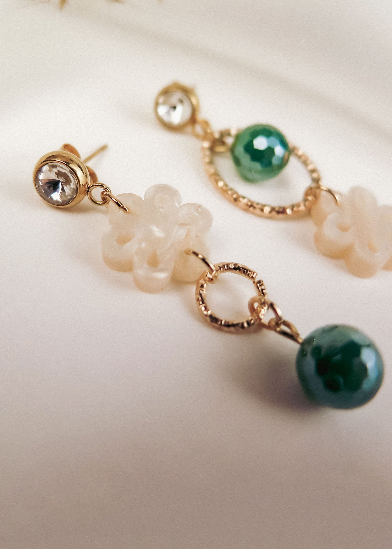 Close-up of two mismatched earrings featuring a clear gemstone stud, a gold ring, a white flower-shaped bead, and a green faceted bead, all laid out on a soft, neutral background for modern charm.
