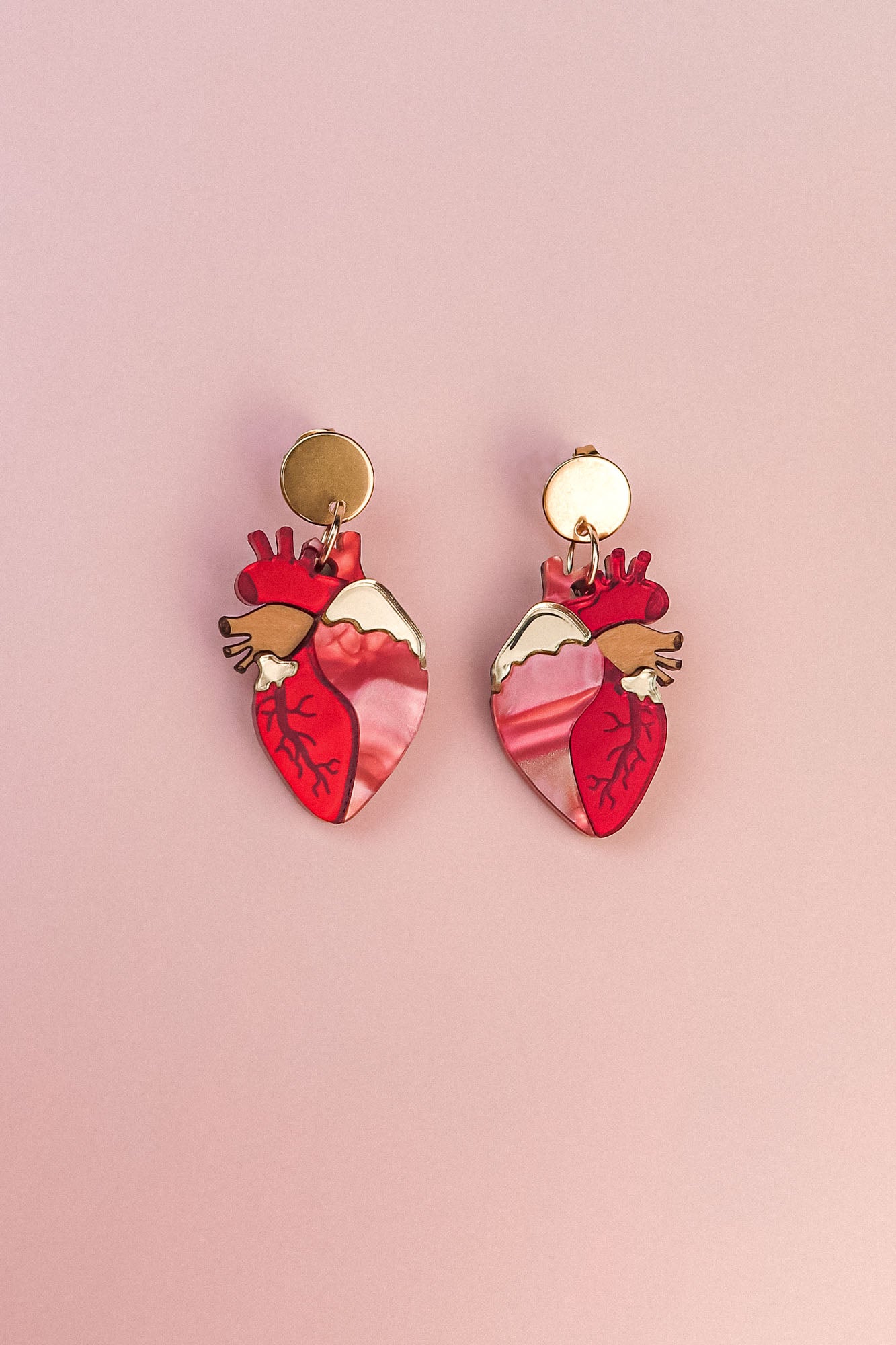 A pair of gold and pink Anatomical Heart Earrings, crafted in acrylic with detailed vein patterns and a unique style, displayed on a light pink background.