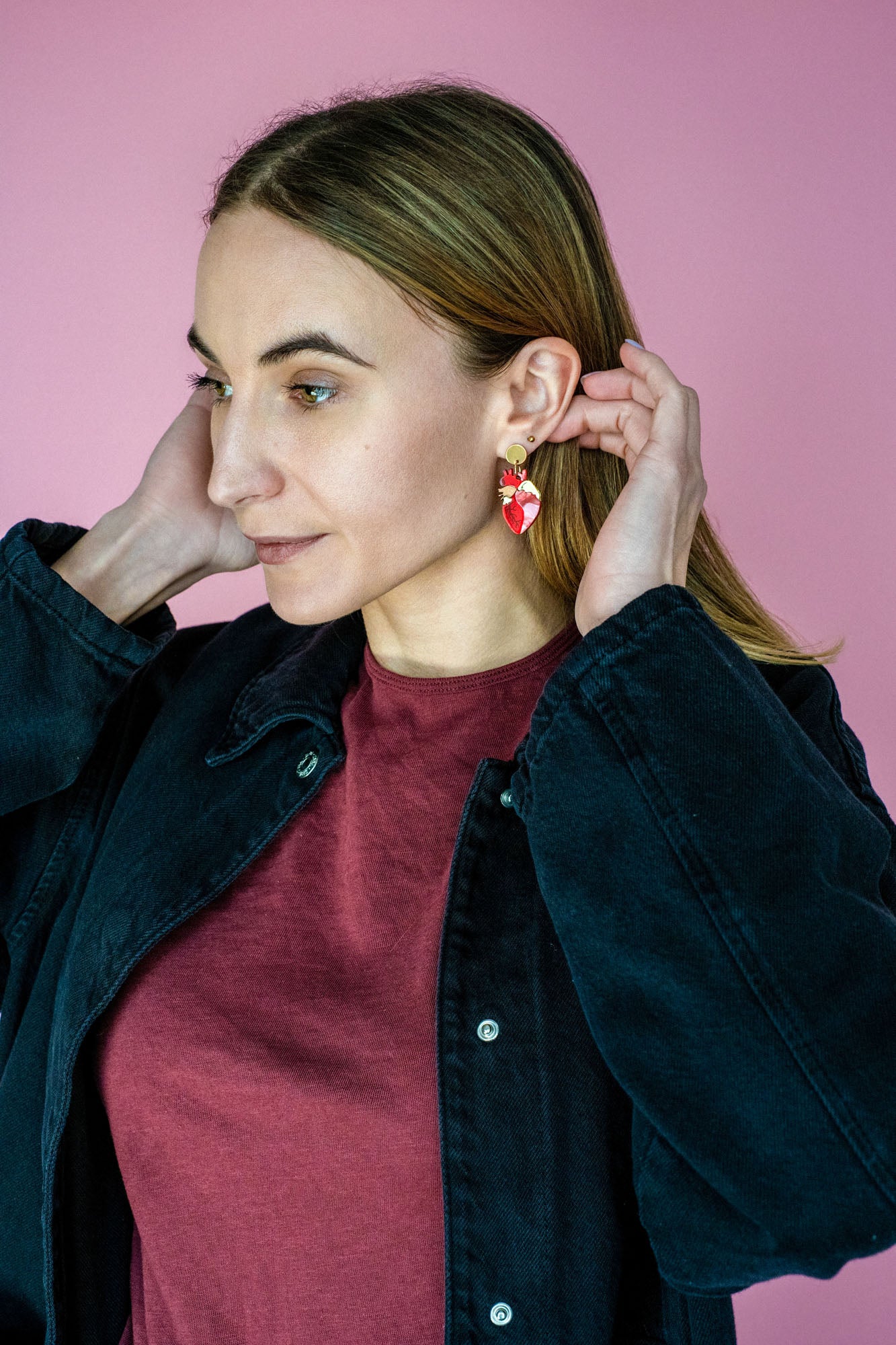 A woman with light brown hair wears a maroon shirt and black jacket, adjusting her unique acrylic earrings. She stands in front of a pink background, looking to the side with a slight smile.