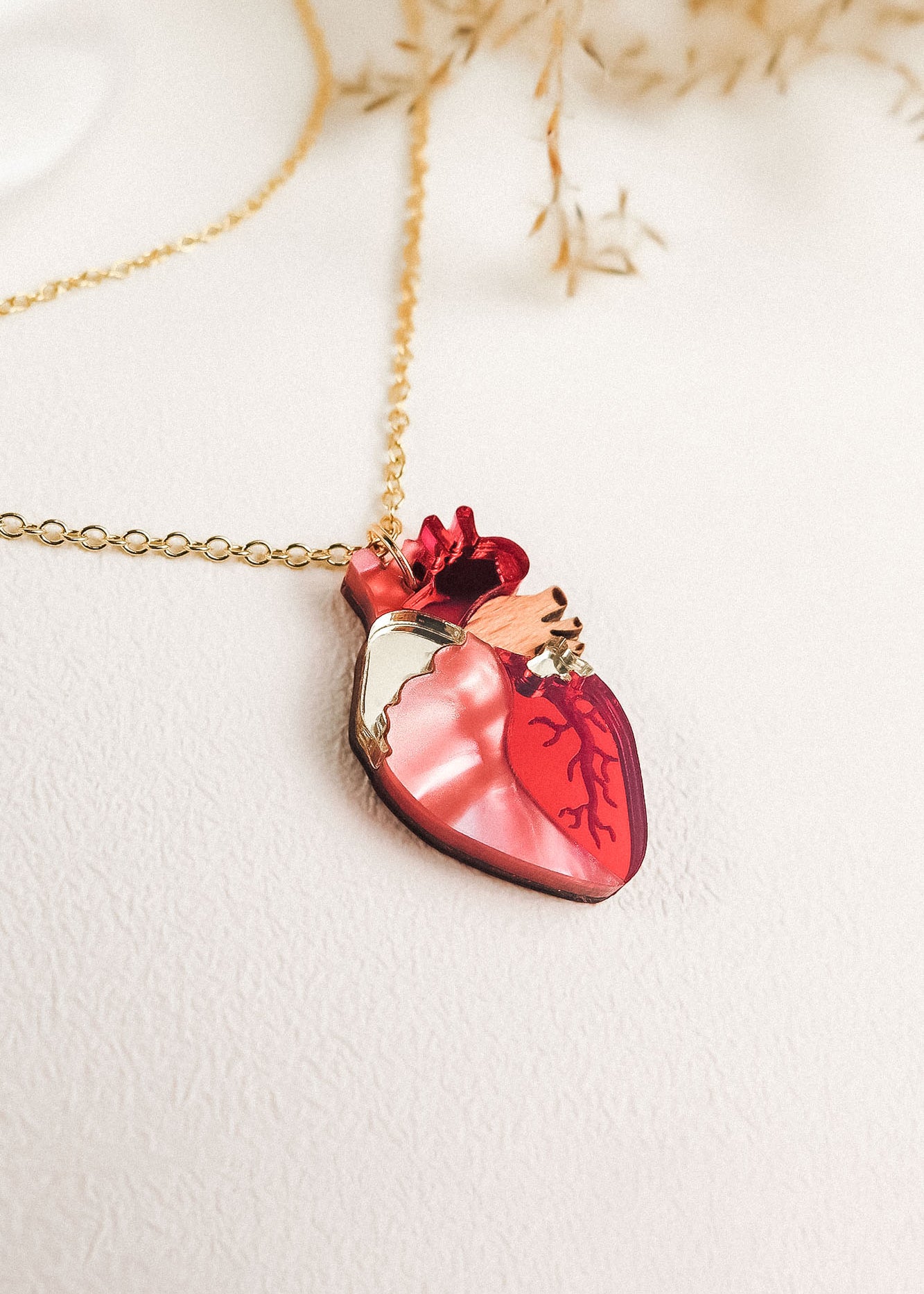 A gold chain anatomical heart necklace with a pendant in red and pink tones, resting on a light textured surface with dried plant accents—a unique jewelry piece that stands out.