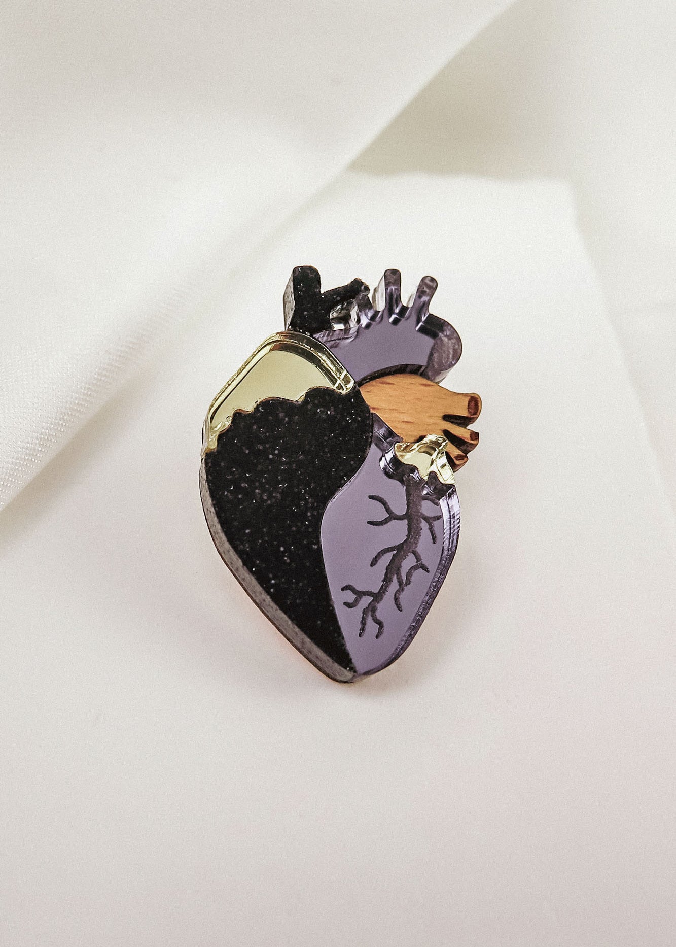 A handcrafted Anatomical Heart Pin, this decorative enamel statement piece features black, gold, and purple sections with detailed veins, beautifully displayed on a white fabric background.