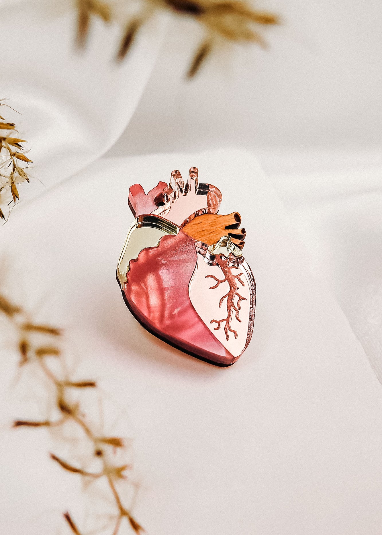 A unique handcrafted glass object shaped like an anatomical heart, featuring shades of pink and gold, sits on a soft white surface with blurred dried plant stems in the foreground.