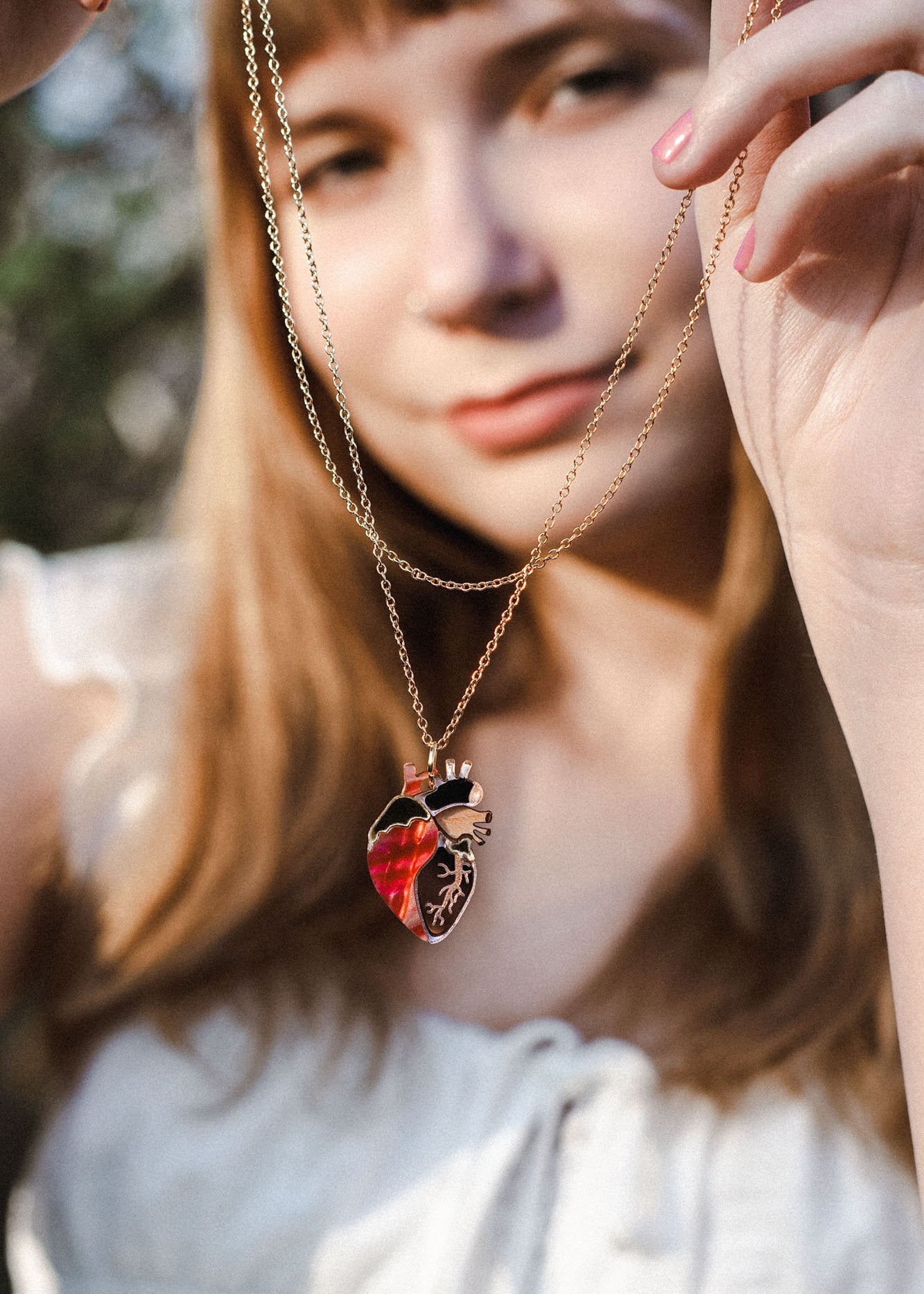 A woman in a white blouse holds up an anatomical heart necklace, the gold pendant in sharp focus while her face remains softly blurred, highlighting this piece of unique jewelry.