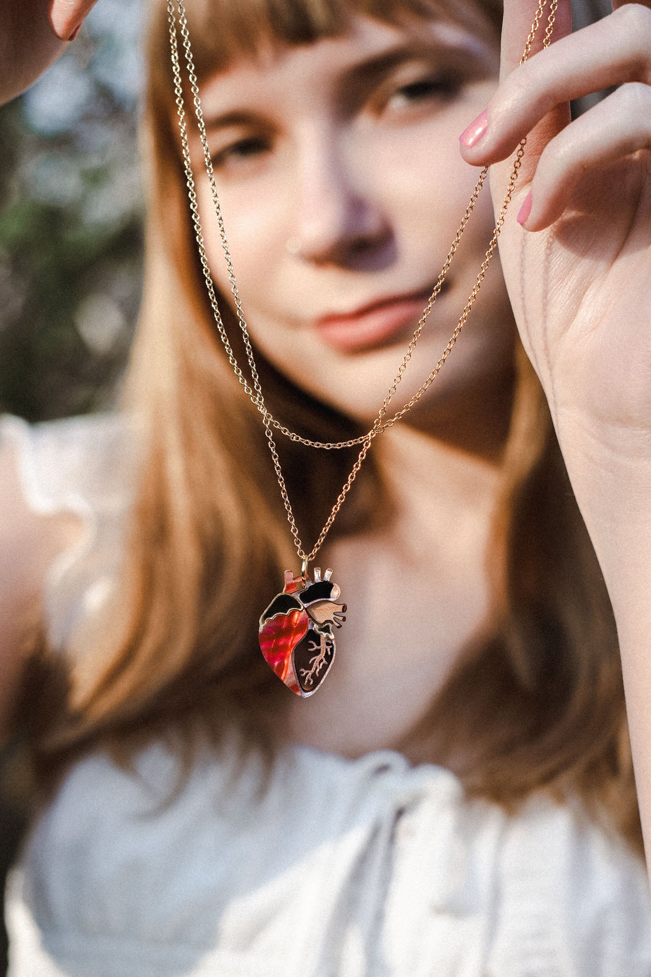 A woman in a white blouse holds up an anatomical heart necklace, the gold pendant in sharp focus while her face remains softly blurred, highlighting this piece of unique jewelry.