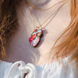 Close-up of a person wearing an anatomical heart necklace in red, pink, and white—a striking piece of unique jewelry. The handcrafted pendant hangs over a white top with a lace-up detail.