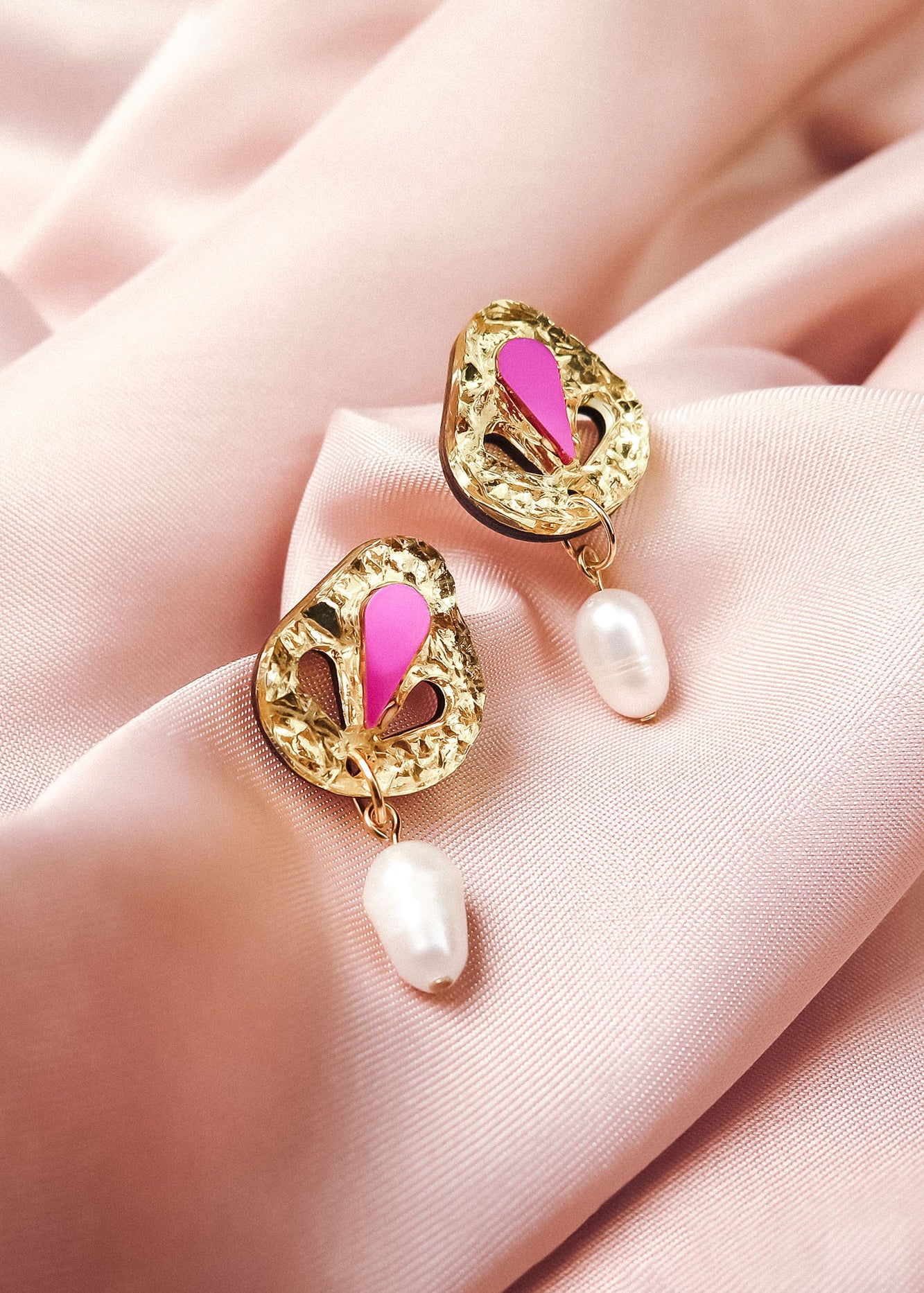 A pair of handcrafted gold drop earrings with intricate designs, pink enamel accents, and white pearl drops, beautifully displayed on soft, pale pink fabric.