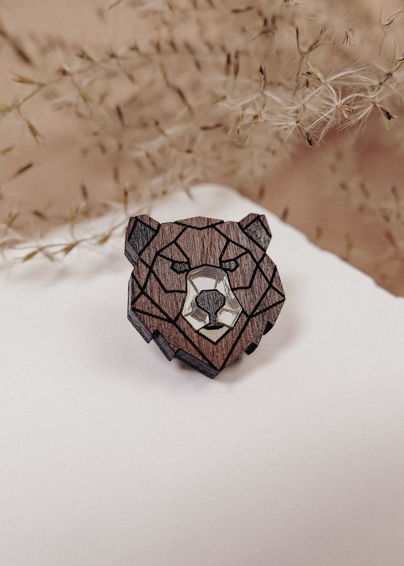A handcrafted wooden brooch in geometric style, shaped like a bear’s head, rests on a white surface with delicate dried plants softly blurred in the background.