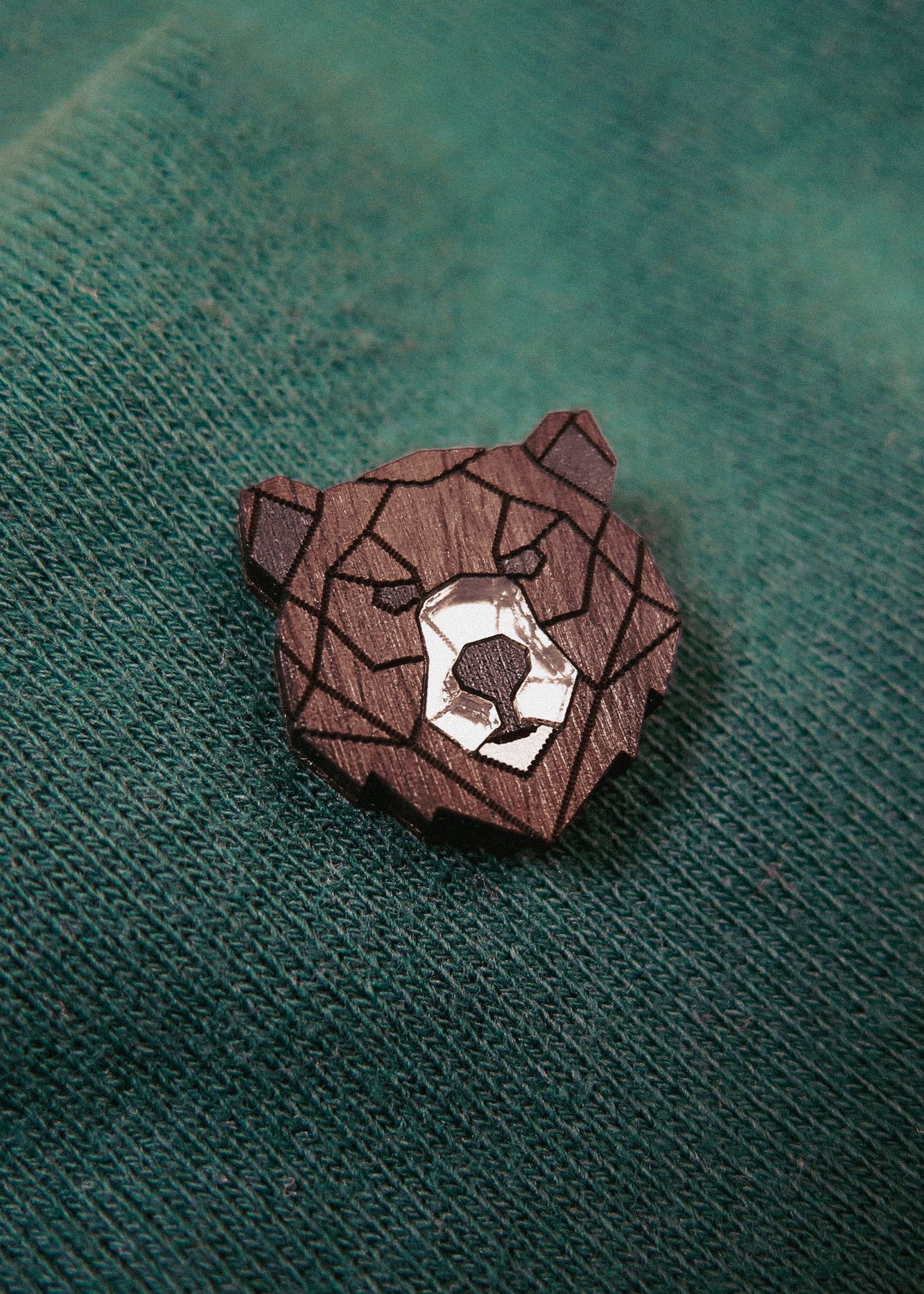 A handcrafted wooden brooch in a geometric style, shaped like a bear’s head, is pinned to green textured fabric. The brooch features angular lines and a white accent on the bear’s snout.