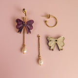 Three mismatched earrings on a pink background: a gold-plated hoop earring, a purple bow with a dangling freshwater pearl, and a white bow edged in gold attached to a bar and pearl—perfect for creating your own interchangeable dangle earring look.