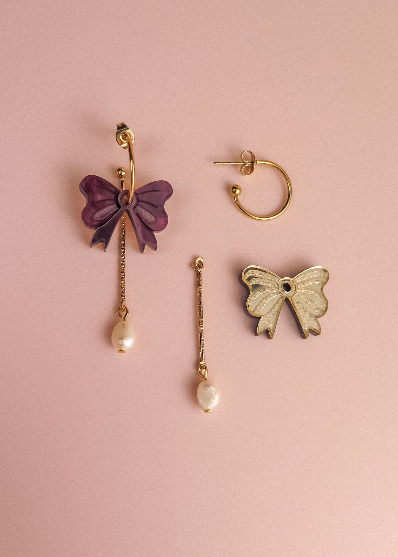 Three mismatched earrings on a pink background: a gold-plated hoop earring, a purple bow with a dangling freshwater pearl, and a white bow edged in gold attached to a bar and pearl—perfect for creating your own interchangeable dangle earring look.
