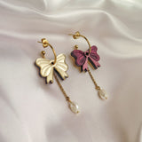 Two gold-plated hoop earrings with bow-shaped charms—one cream and one pink—each with a dangling gold chain and a freshwater pearl, displayed on white fabric with decorative gold spheres.