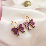 A pair of gold-plated hoop earrings with purple and pink butterfly-shaped charms are displayed on white satin fabric, accented by a pink ribbon and gold decorative balls nearby.