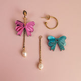 Three mismatched interchangeable dangle earrings on a pink surface: a gold-plated c-hoop, a dangling earring with a pink bow and freshwater pearl, and another with a blue bow, sequins, and pearl.
