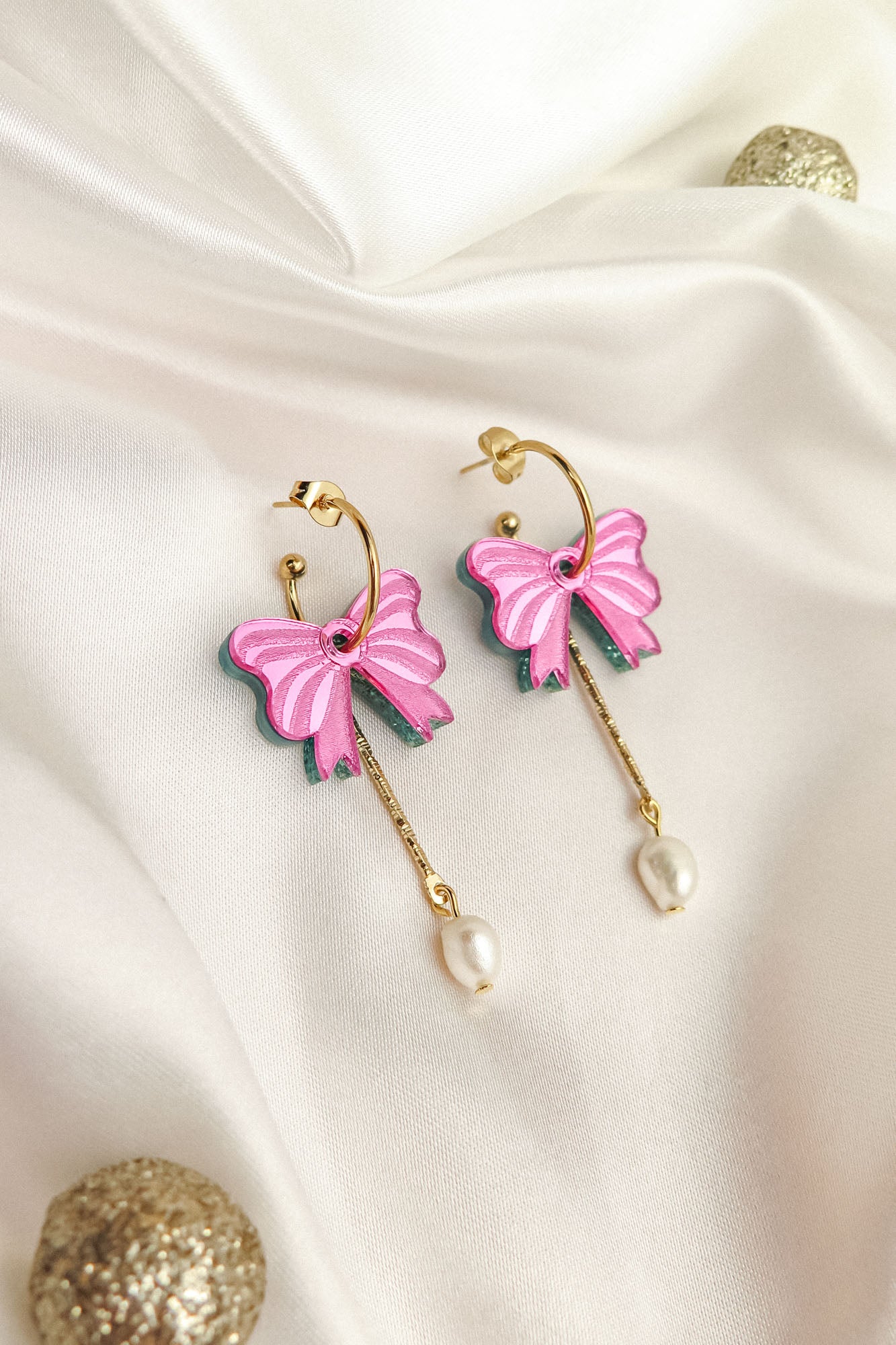 A pair of gold-plated c-hoop earrings with pink butterfly charms and freshwater pearl drops, displayed on white satin fabric with decorative gold spheres nearby.