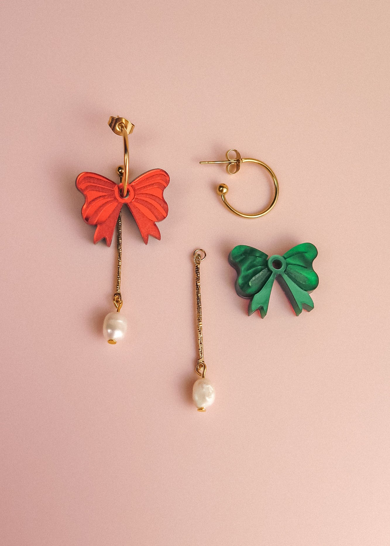 A pair of mismatched earrings on a pink background: one features a red bow, gold bar, and freshwater pearl; the other showcases a green bow with a gold-plated c-hoop, gold bar, and pearl for an interchangeable dangle earrings look.