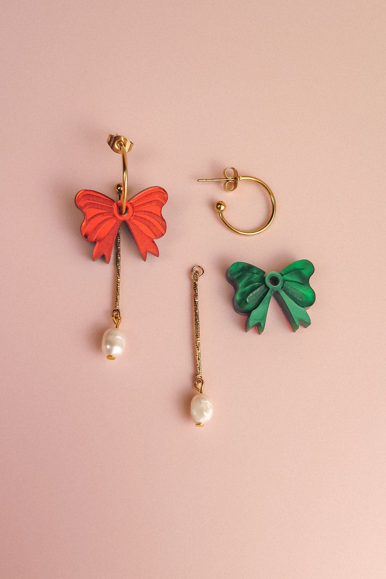 A pair of mismatched earrings on a pink background: one features a red bow, gold bar, and freshwater pearl; the other showcases a green bow with a gold-plated c-hoop, gold bar, and pearl for an interchangeable dangle earrings look.