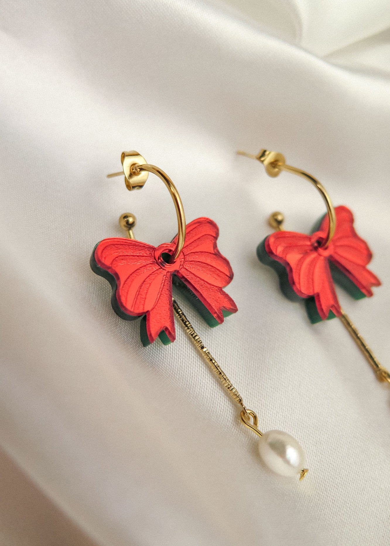 A pair of gold-plated c-hoop earrings with red butterfly-shaped charms and dangling gold chains ending in a single freshwater pearl, displayed on white satin fabric.