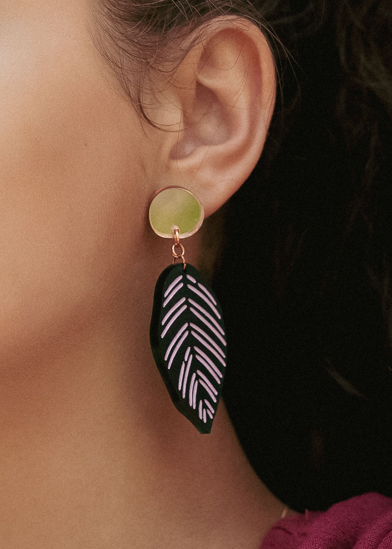 Close-up of a persons ear wearing Calathea Ornata Earrings—handcrafted earrings with a circular gold stud and a black leaf-shaped charm adorned with pink geometric patterns, inspired by tropical nature jewelry. The person has dark hair and wears a maroon top.