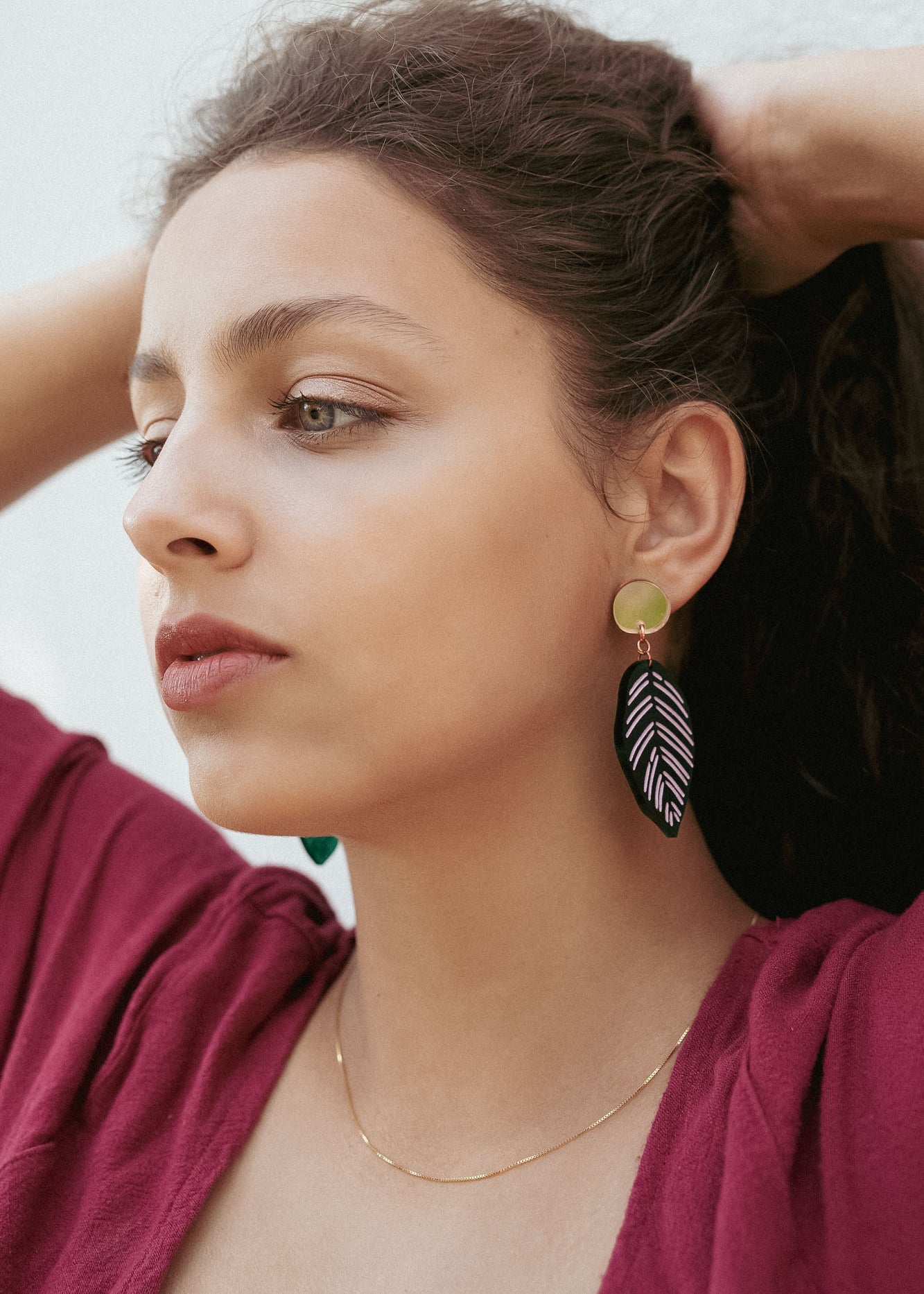 A woman with wavy brown hair pulled back wears a burgundy top, a delicate gold necklace, and Calathea Ornata Earrings—handcrafted earrings inspired by tropical nature jewelry—featuring green beads and a black-and-white leaf design.