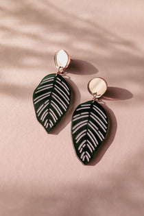 A pair of handcrafted earrings with round gold studs and green leaf-shaped pendants with white vein details, inspired by Calathea Ornata Earrings, displayed on a pink surface with soft shadows.