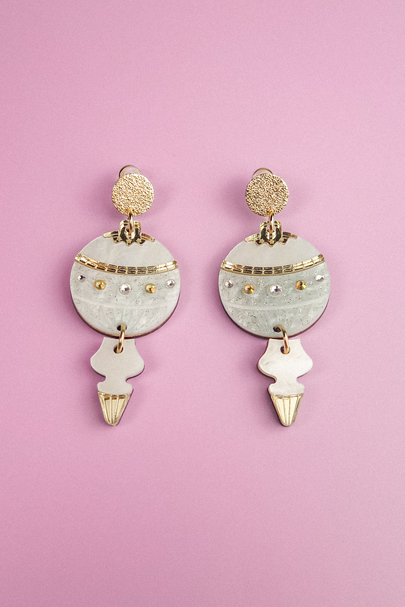 A pair of ornate, gold and white festive earrings with circular and abstract shapes, resting on a pastel purple background.