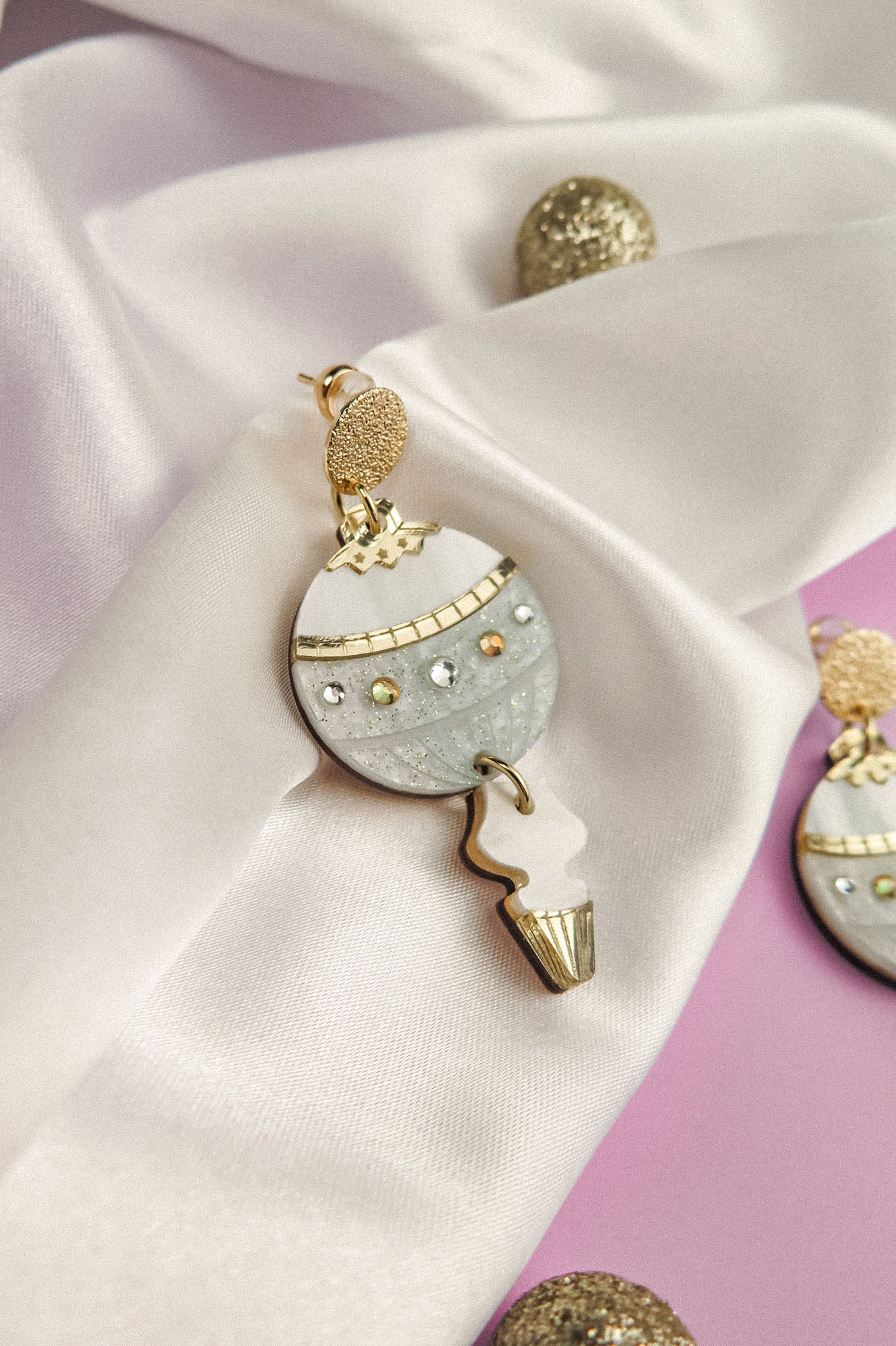 A decorative festive earring with a round, silver and gold design, studded with gems, rests on white satin fabric with gold and glitter accents. The perfect holiday jewelry piece, set against a soft lavender background.