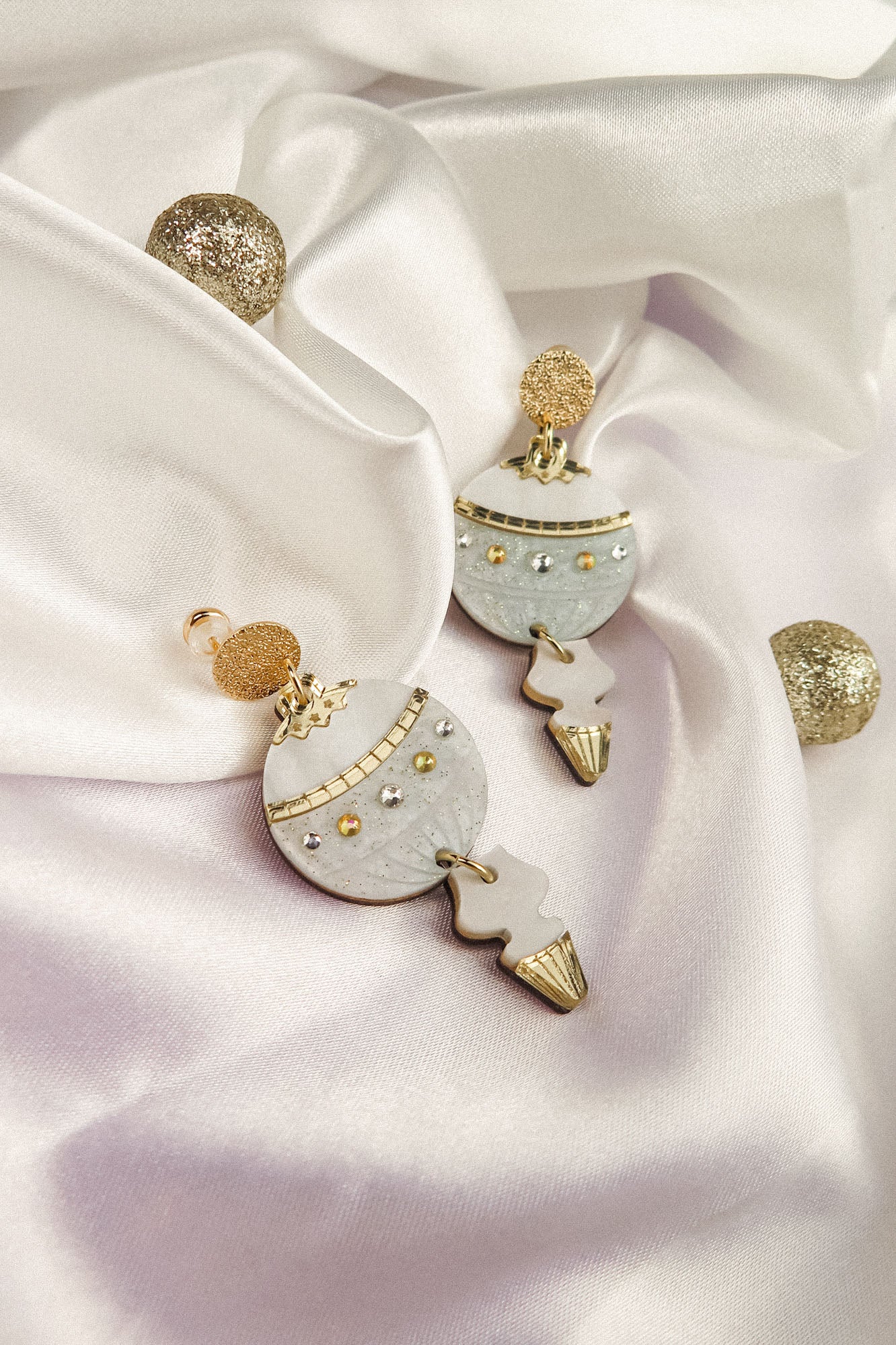 A pair of decorative Poinsettia Statement Hoops with gold, white, and silver details are displayed on white satin fabric, accompanied by glittery gold spheres—perfect as festive earrings to elevate your holiday jewelry collection.
