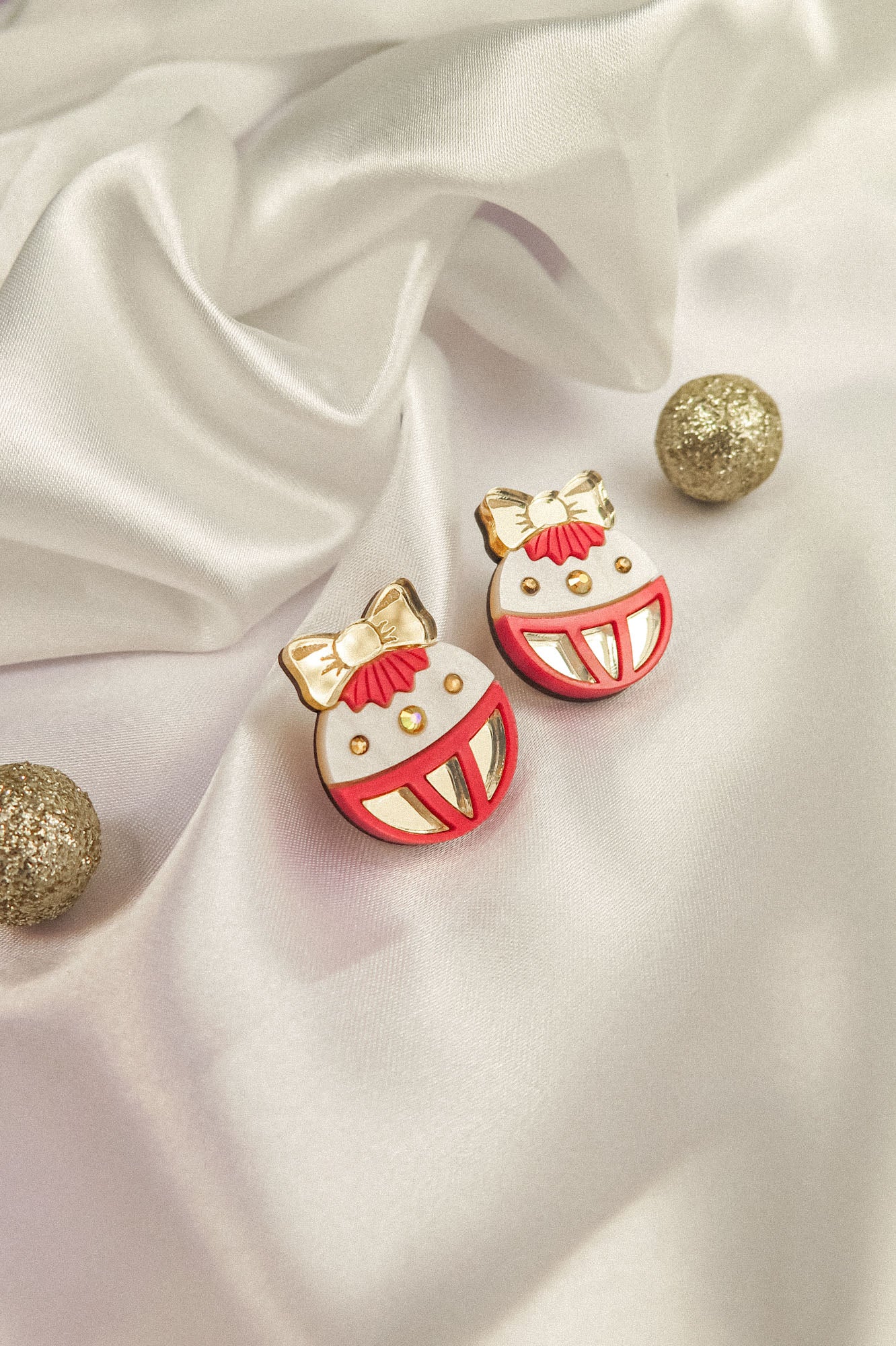 Two round, decorative festive earrings with red, white, and gold details, featuring bows and smiling faces, are placed on white satin fabric amid three glittery gold spheres—perfect as holiday jewelry or Poinsettia Statement Hoops.