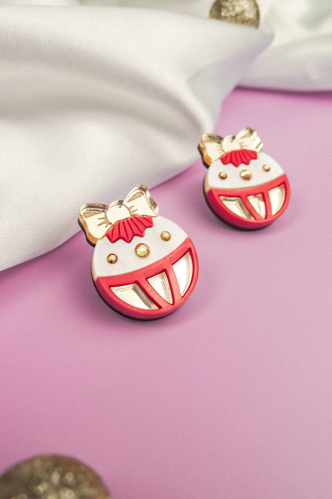 A pair of colorful enamel pins shaped like smiling faces with big red mouths, gold bows, and gold accents, bring a touch of festive earrings style, placed on a purple surface next to white fabric.