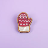 A Christmas cookie shaped like a mitten, inspired by a Christmas Mitten Pin, is decorated with red, white, and pink icing and lies on a light purple background for a festive holiday style.