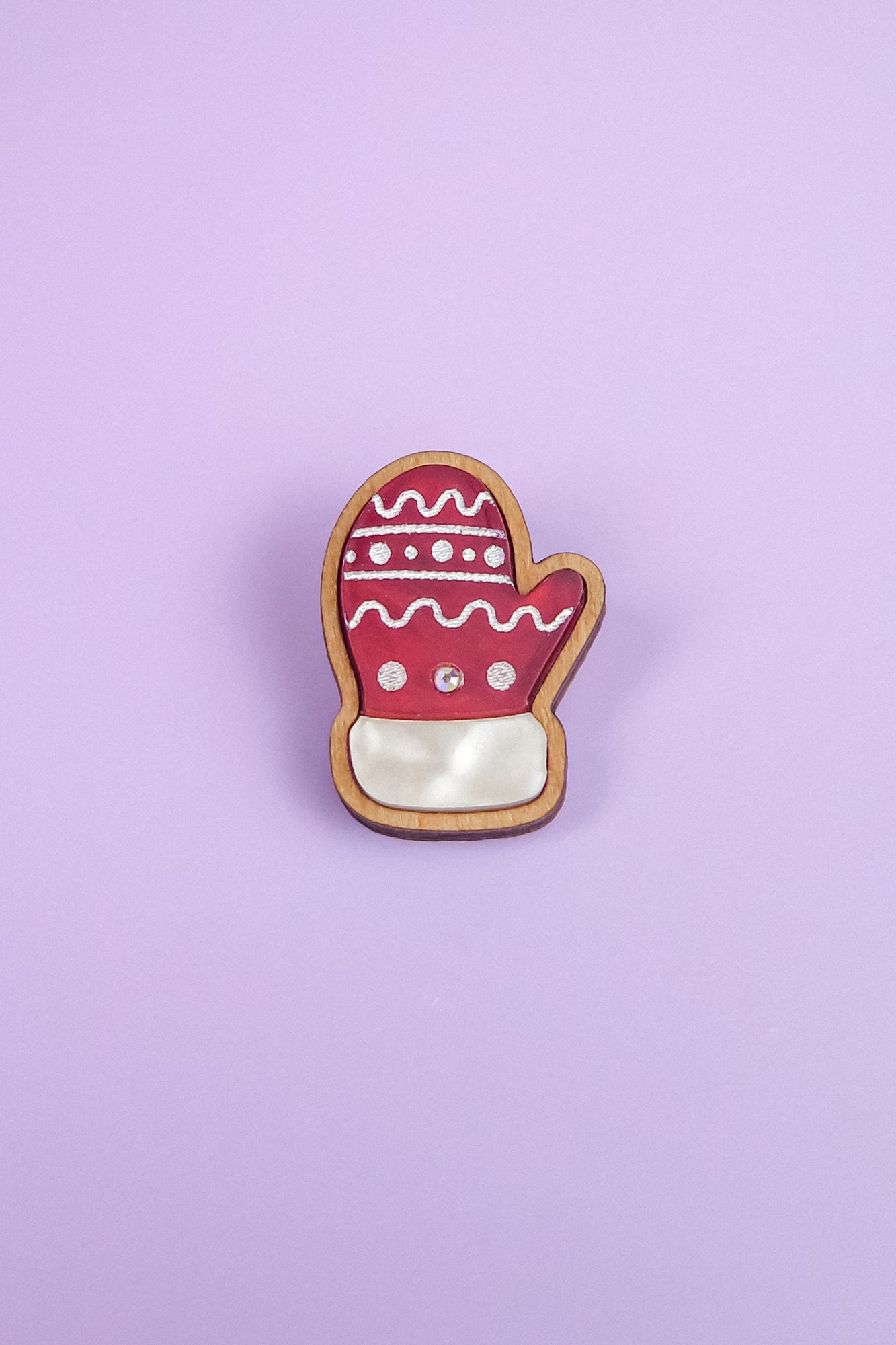 A Christmas cookie shaped like a mitten, inspired by a Christmas Mitten Pin, is decorated with red, white, and pink icing and lies on a light purple background for a festive holiday style.