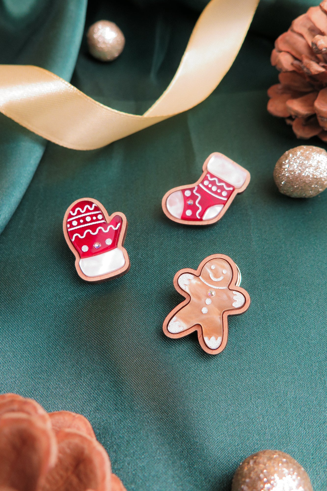 Three decorated cookies shaped like a mitten, a stocking, and a gingerbread person rest on green fabric with gold ribbon, glittery ornaments, pinecones, and a handcrafted accessory to complete the festive holiday style.