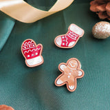 Three handcrafted Christmas cookies—a mitten, a stocking, and a gingerbread person—sit on green fabric with a gold ribbon, pinecones, and sparkly ornaments, adding festive charm to your holiday wardrobe.