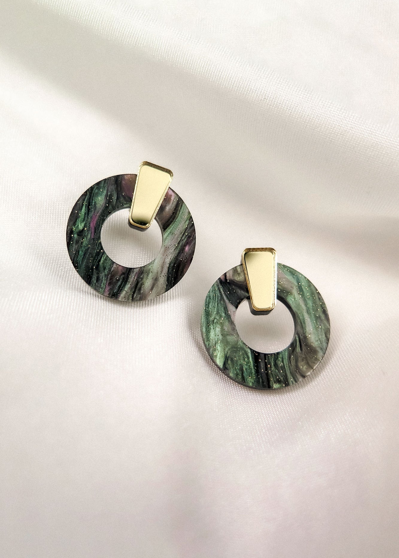 A pair of round, marbled green and black acrylic glass earrings with gold triangular studs, displayed on a soft white fabric background. These hypoallergenic Minimalist Circle Stud Earrings combine style and comfort beautifully.
