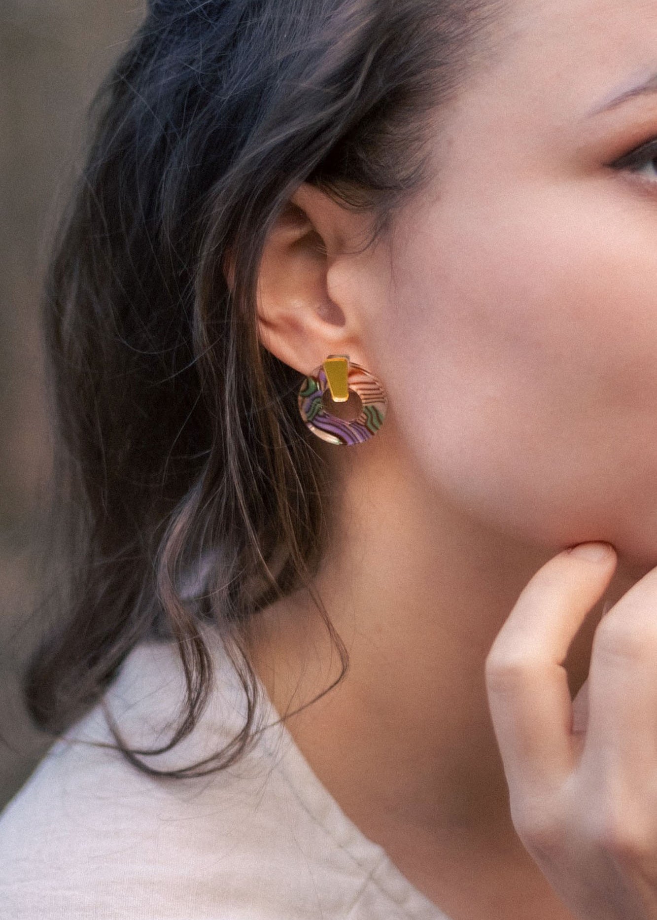A close-up of a woman wearing minimalist circle stud earrings crafted from acrylic glass, featuring abstract multicolored patterns and a gold rectangular accent. She has dark hair and is touching her chin with her hand.