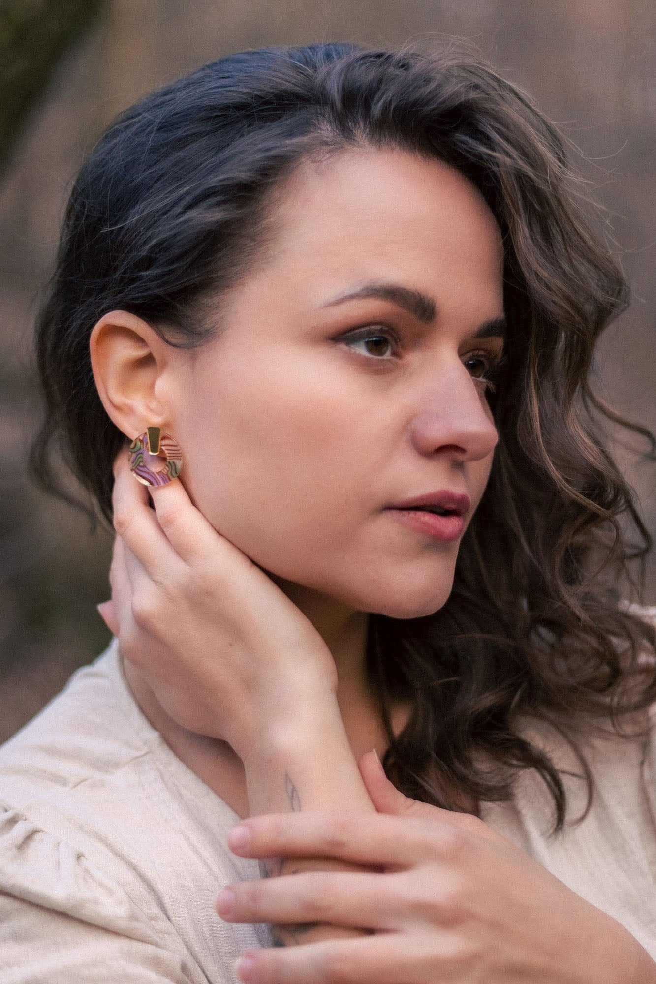 A woman with wavy brown hair wearing a beige top touches her Minimalist Circle Stud Earrings with one hand and rests her other hand on her collarbone. She looks slightly to the side with a calm expression. The earrings are hypoallergenic.