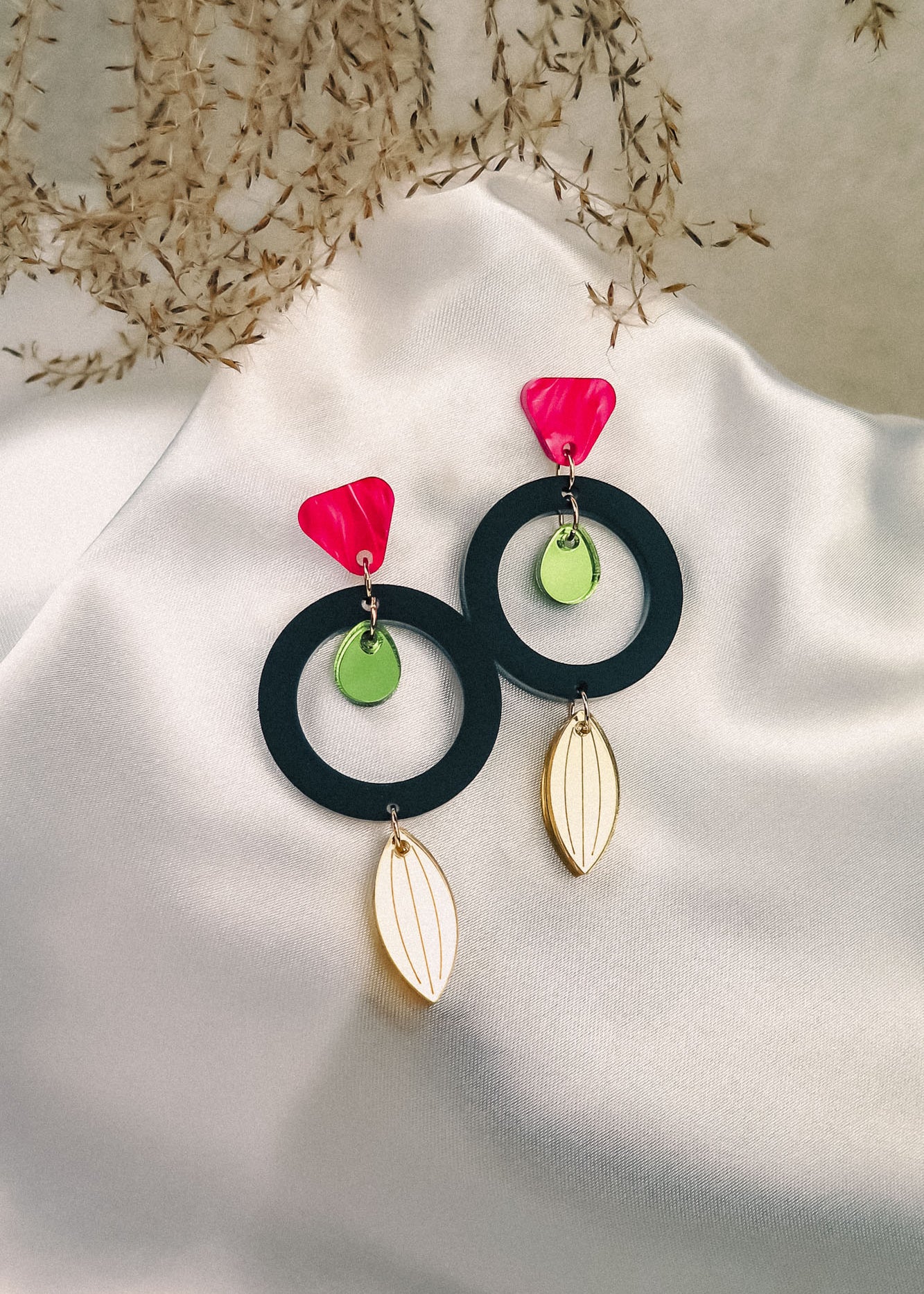 A pair of modern, colorful dangle earrings crafted from recycled acrylic, featuring pink triangle studs, green circular accents, black hoops, and gold leaf-shaped dangles displayed on white satin with dried foliage in the background.