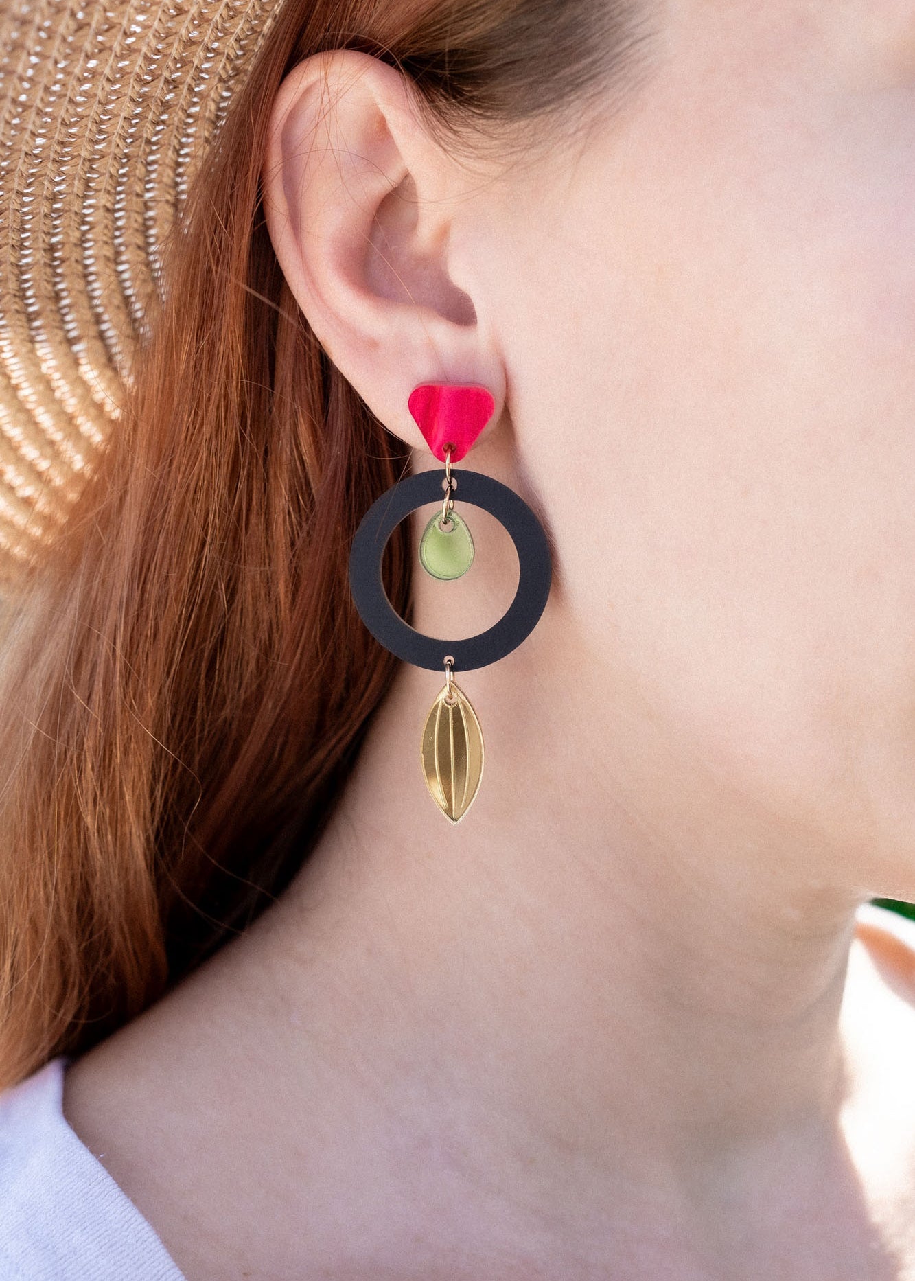 A woman in a straw hat and white shirt showcases artisan jewelry: dangle earrings crafted from recycled acrylic, featuring a black circle, gold shapes, and a red accent near the ear for a striking statement look.