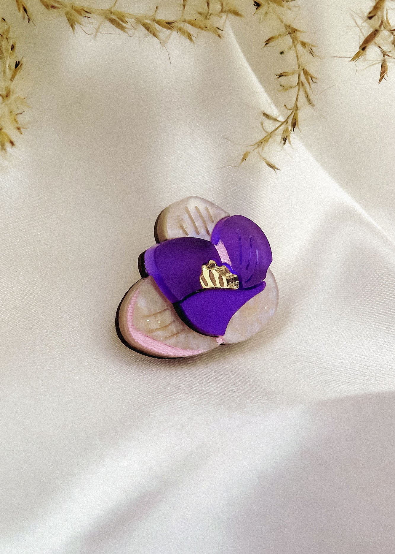 A purple and cream flower-shaped brooch, reminiscent of a Purple Crocus Flower Lapel Pin with gold detailing, rests on smooth white satin fabric, surrounded by delicate dried grass—perfect for nature lovers.