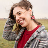 A smiling woman with brown hair stands outdoors in a grassy field, wearing a red sweater, a plaid coat, and festive Poinsettia Statement Hoops. She holds her hair back with one hand on a cloudy day, adding a touch of holiday earrings to her look.