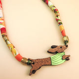 A piece of handcrafted jewelry featuring a colorful, patterned fabric cord and a wooden pendant shaped like a leaping rabbit in a green shirt. Made with recycled materials, set against a soft beige background.