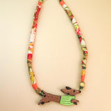 A colorful handcrafted Dachshund necklace featuring a patterned fabric chain and a pendant of a running dachshund in a green sweater. Made with recycled materials, it boasts gold details against a light beige background.