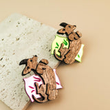 Two playful brooches shaped like reclining capybaras, each resting on a colorful pillow—one pink and one neon green—placed on a textured stone surface. A charming touch for any dog accessory collection or fans of unique brooches.