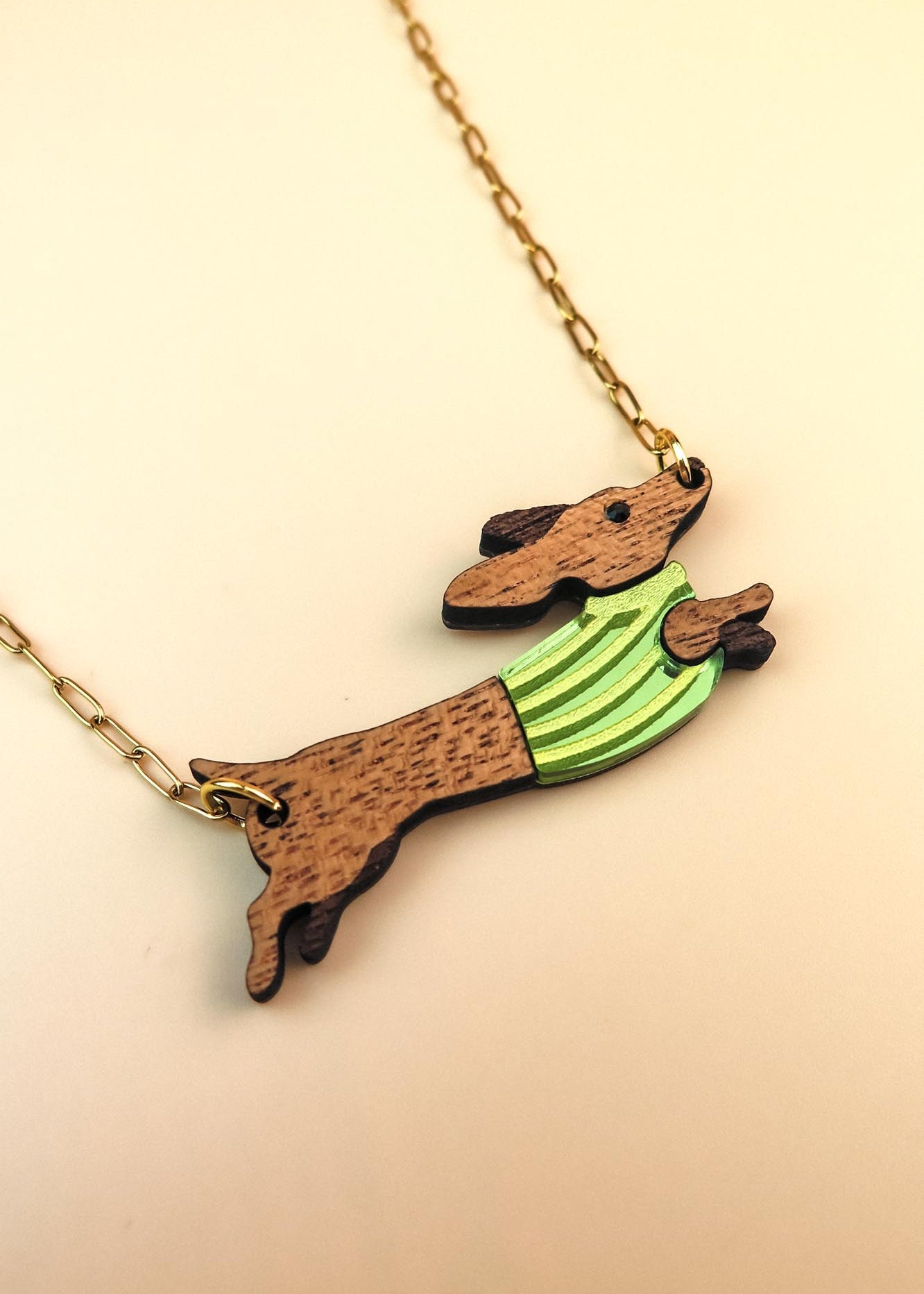 A gold chain necklace featuring a wooden Dachshund pendant in a colorful sweater, crafted from sustainable wood. The leaping pup wears green and yellow stripes against a light cream background.