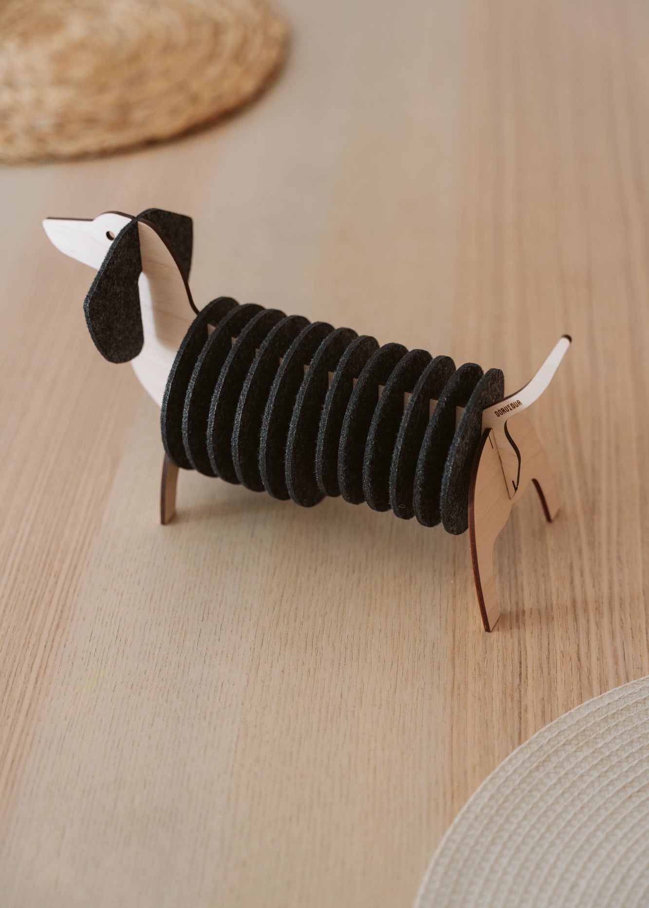 A napkin holder shaped like a dachshund dog, made of wood and black felt, sits on a light wooden table with woven placemats and matching Dachshund coasters visible in the background.