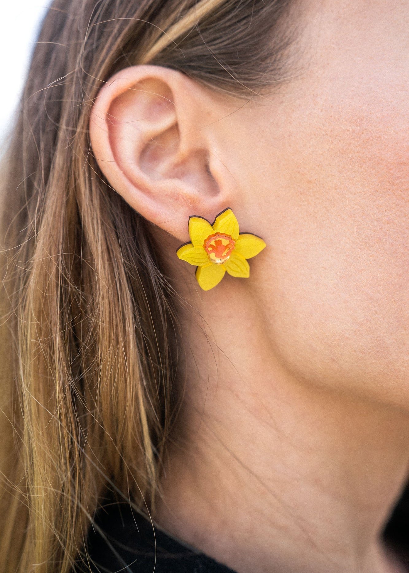 Close-up of a womans ear wearing Daffodil Stud Earrings with an orange center. Her light brown hair is tucked behind her ear, and she is dressed in a black top—perfect unique accessories for stylish women.