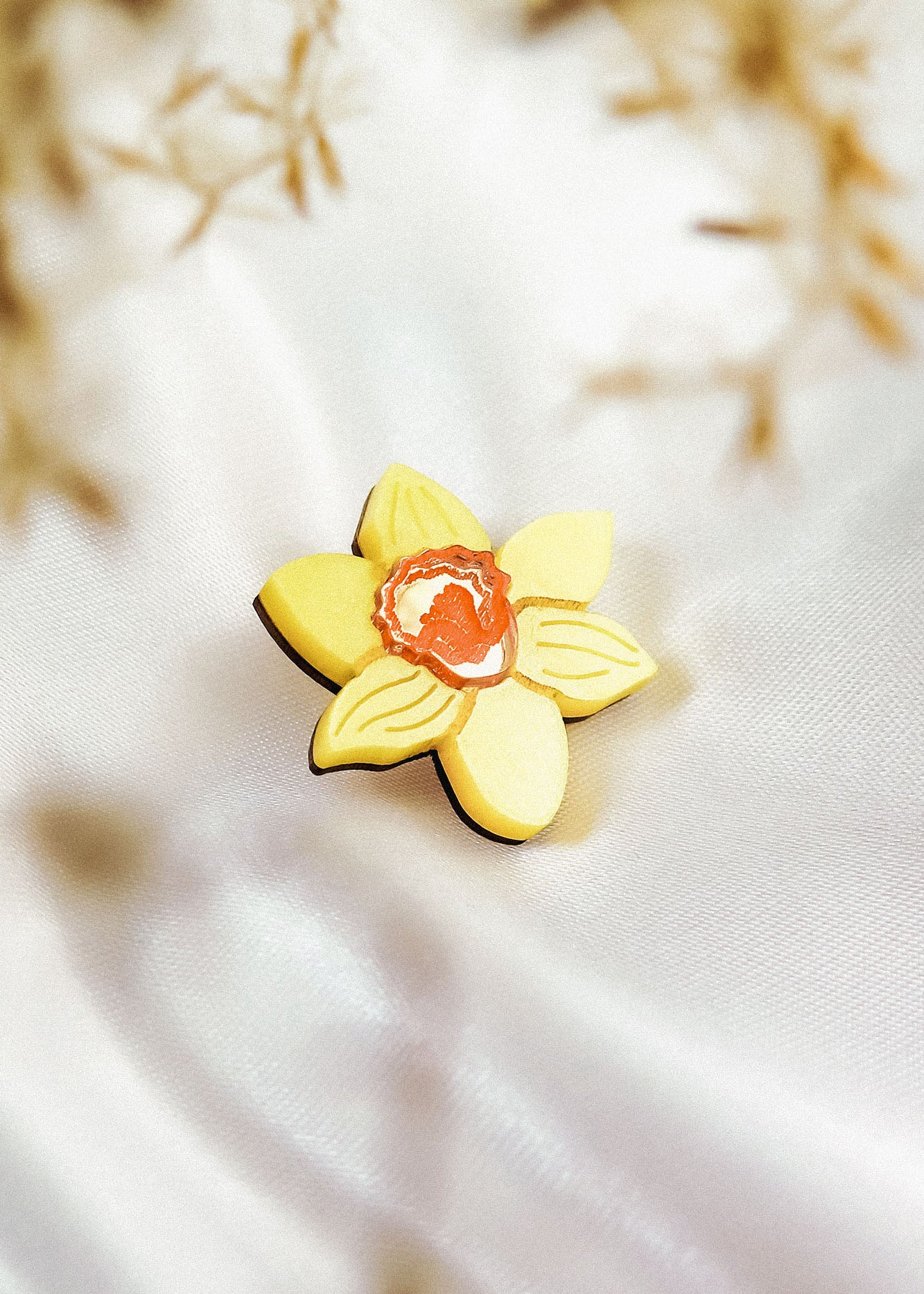 A yellow, handmade flower-shaped cookie with an orange center rests on white, silky fabric, resembling a Daffodil Flower Lapel Pin, with blurred brownish plants in the background.