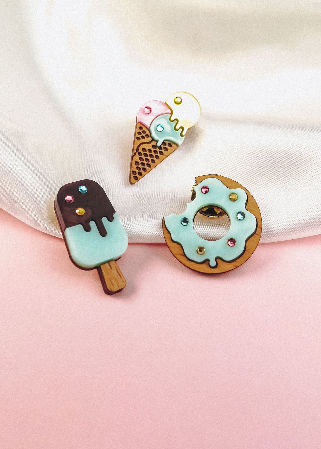 Three wooden pins shaped like desserts—a popsicle, an ice cream cone, and a handcrafted Donut Pin—decorated with pastel icing and colorful sprinkles, are displayed on a pink and white background.