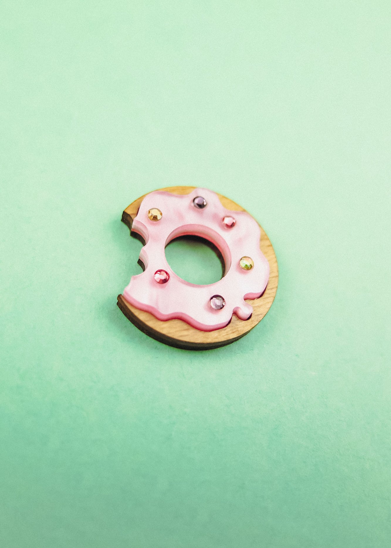 A handcrafted wooden Donut Pin with pink icing and colorful rhinestones, featuring a bite mark, sits on a mint green background.
